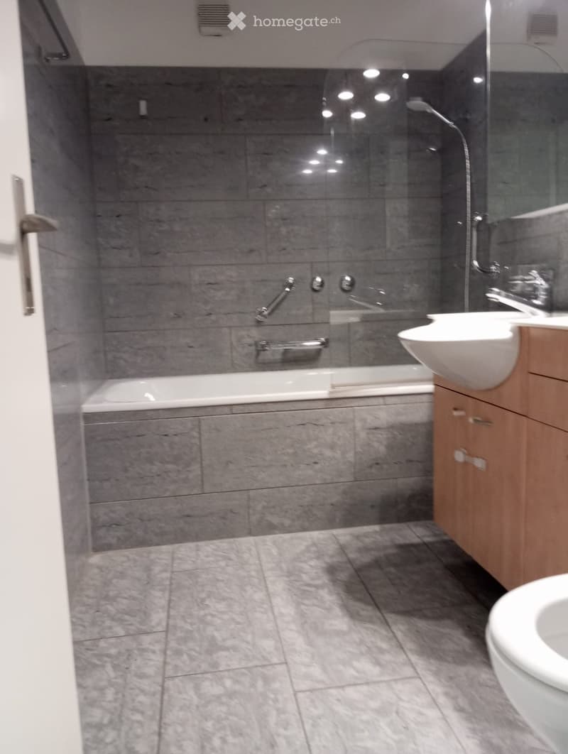 Modern bathroom with grey tiled walls and floor, featuring a bathtub, sink, toilet, and wooden cabinet.