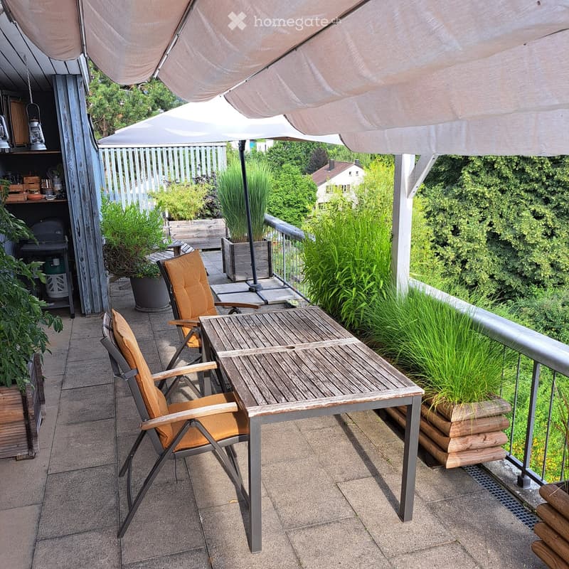 Balcony with a wooden table, two chairs, potted plants, and a large umbrella providing shade.