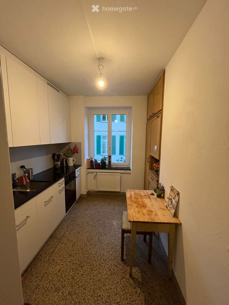modern kitchen with white cabinets, black countertops, wooden table, small window, radiator, wooden chair, small sink