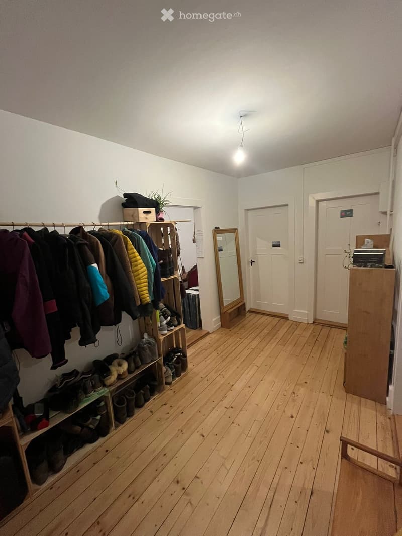 Empty room with wooden floor, white walls, wooden bench, wooden shelves with clothes and shoes, mirror, and a plant on top of a shelf.