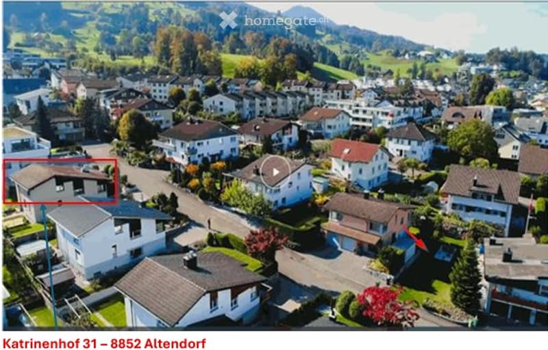Aerial view of a residential neighborhood with multiple houses, surrounded by green fields and mountains in the background.