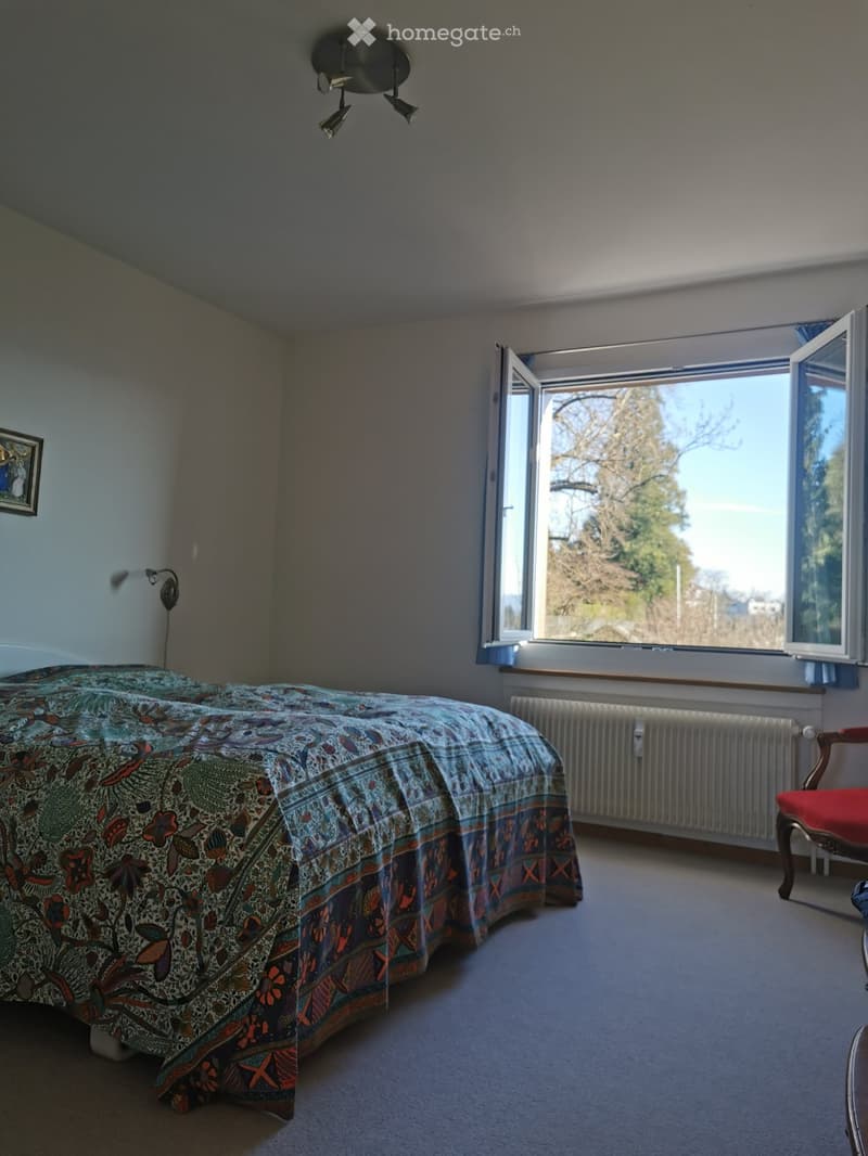 Bedroom with a double bed, colorful patterned duvet, a red chair, a radiator, and a large window with a view of trees.