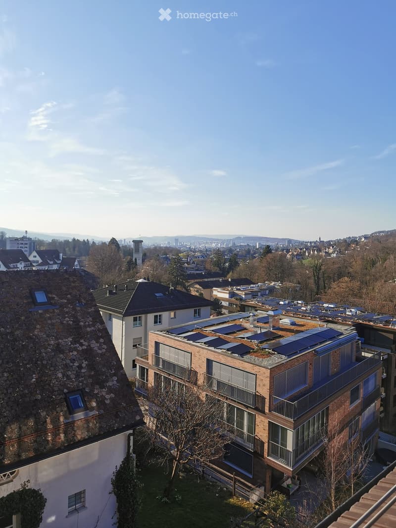 A modern multi-story residential building with solar panels on the roof, surrounded by trees and other houses in a suburban area.