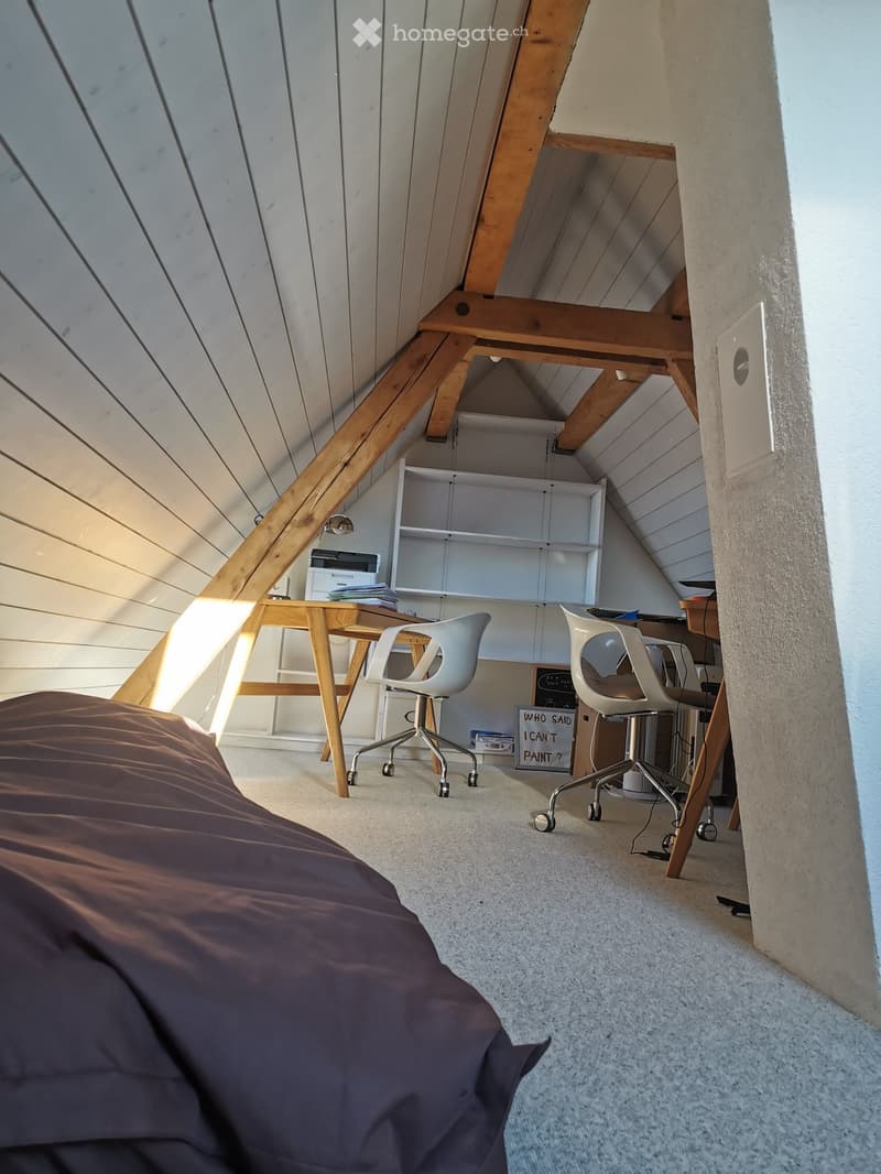 Loft-style room with a slanted wooden ceiling, a bed, a desk with chairs, a shelf, and a printer. Natural light streams in from the left side.