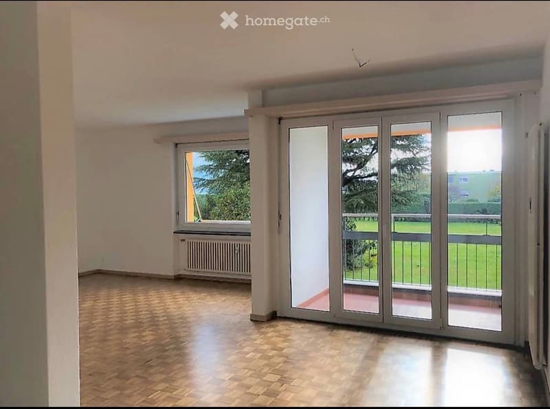 Empty room with wooden flooring, large sliding glass doors leading to a balcony, and a window with a view of greenery.