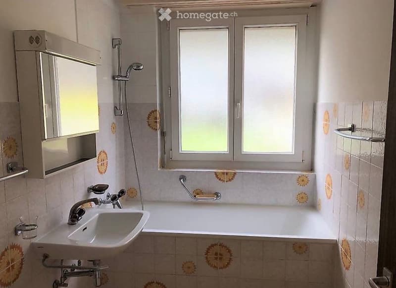 Bathroom with a white sink, bathtub, mirror, and showerhead. The walls are tiled with a floral pattern, and there is a window above the bathtub.