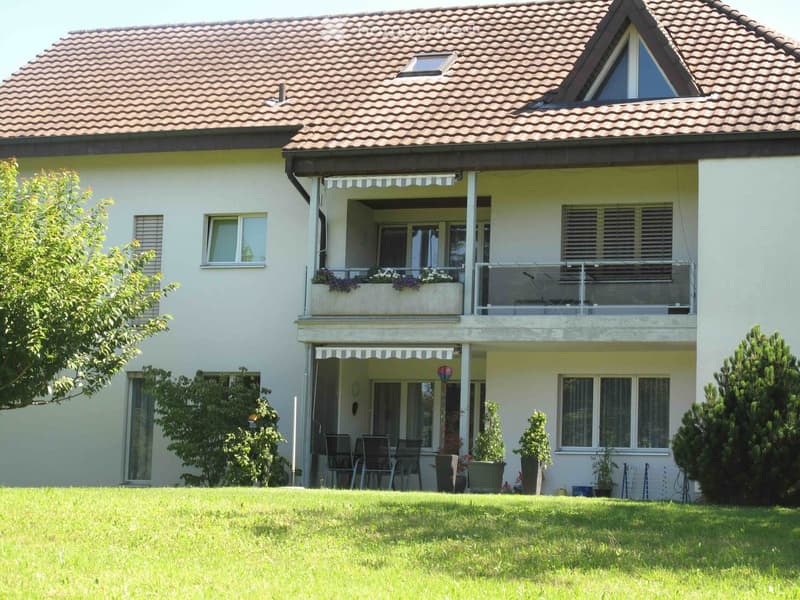 Two-story house with a tiled roof, white exterior, multiple windows, and a covered balcony area with outdoor furniture.