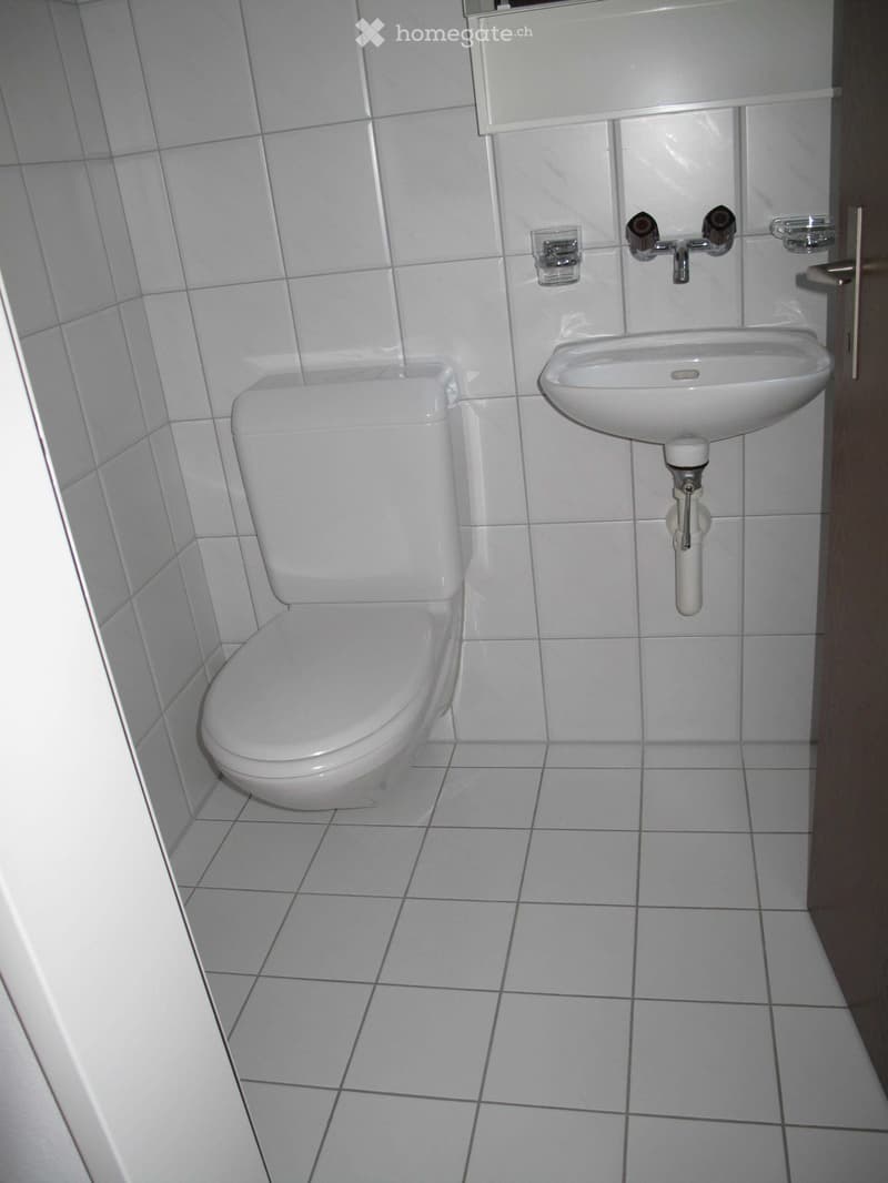 A small bathroom with white tiled walls and floor, featuring a toilet and a sink with a faucet.