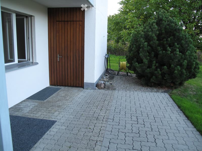 A paved pathway leads to a wooden front door of a house, with a garden area featuring a large bush and a hose reel nearby.