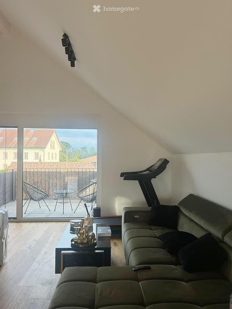 Modern living room with wooden floor, large window offering a view, sofa, coffee table, small table with chairs, and a treadmill in the corner.