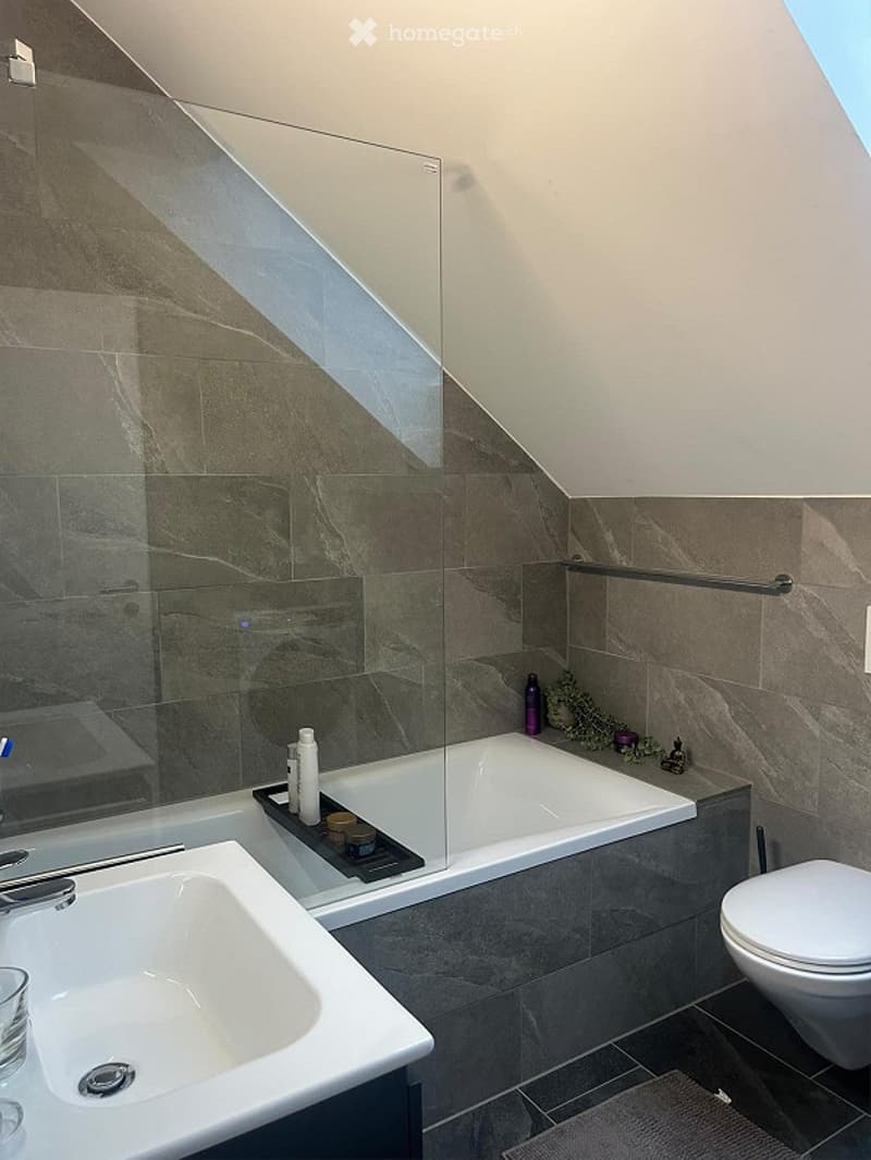 Modern bathroom with a bathtub, toilet, sink, and a glass-enclosed shower area. The walls are tiled in a dark, marble-like pattern, and there is a small tray with toiletries on the edge of the bathtub.