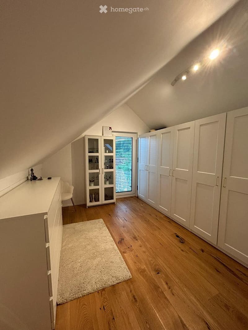 Attic room with wooden flooring, white built-in wardrobes, a small window with blinds, a white cabinet with glass doors, a chair, and a small rug.