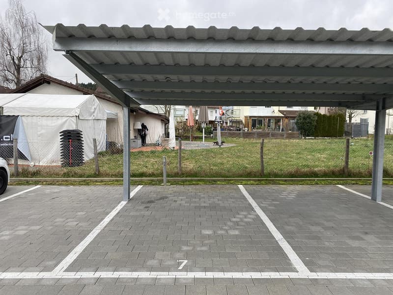 Covered parking area with numbered spaces, adjacent grassy area, and nearby buildings.