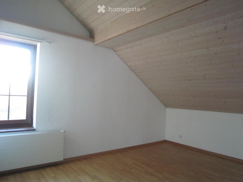 Empty attic room with wooden floor, white walls, and a slanted wooden ceiling. Features a single window and a radiator.