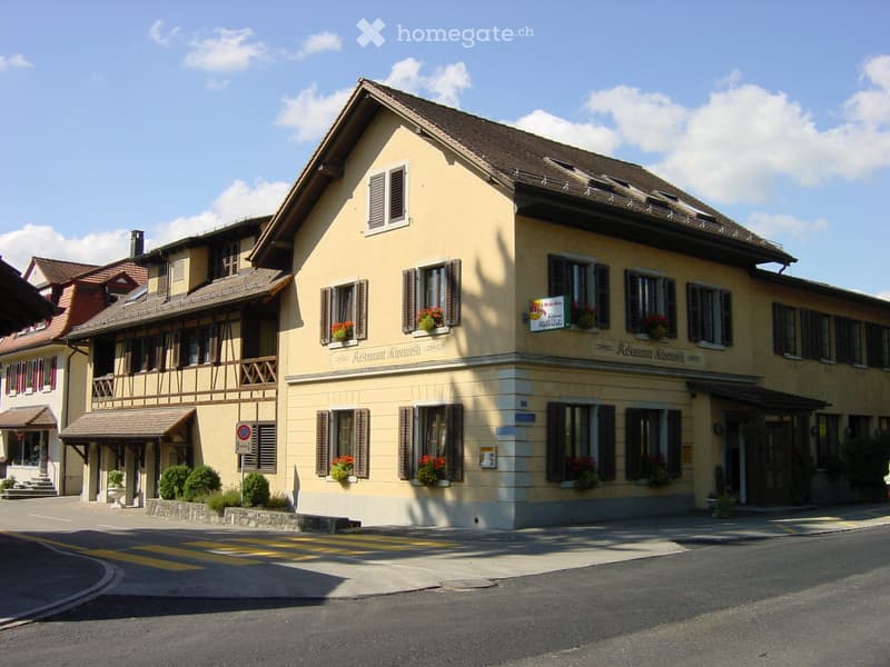 Two-story yellow building with brown shutters, multiple windows, and flower boxes. The building has a sign reading "Restaurant Alveari" and a small balcony on the left side.