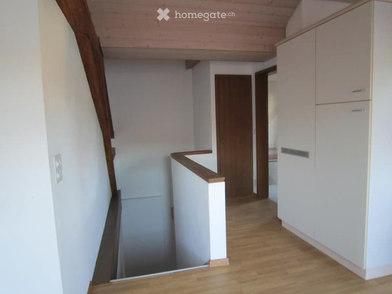 A narrow hallway with wooden flooring, white walls, and a wooden staircase leading upwards. There are white cabinets on the right side and a closed wooden door at the end of the hallway.
