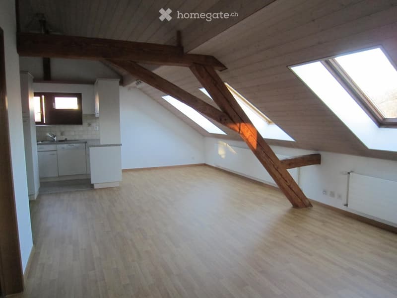 Empty attic space with wooden beams, skylights, and a small kitchen area visible in the background.