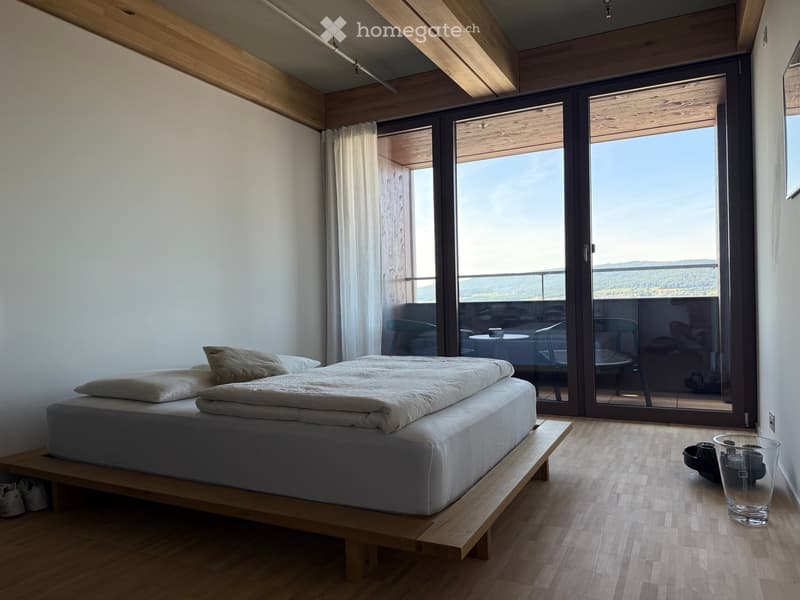 Modern bedroom with a raised wooden bed, large windows offering a scenic view, and a small seating area visible through the glass doors.