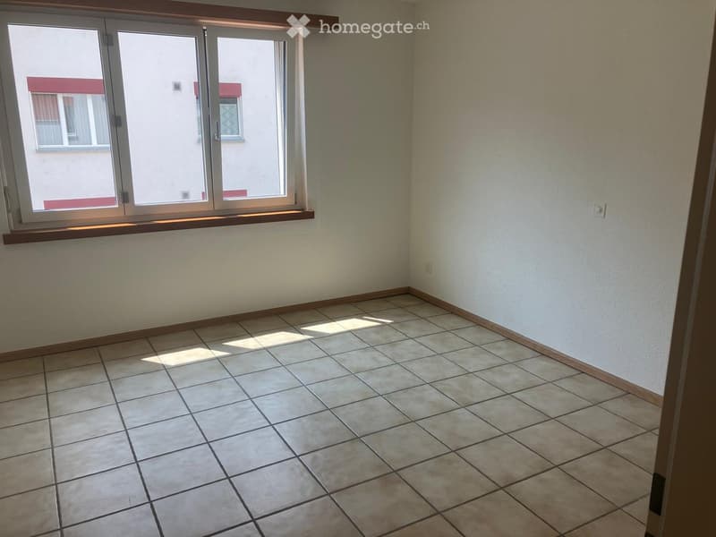 Empty room with tiled floor and large window with wooden sill, showing natural light and a view of neighboring building.