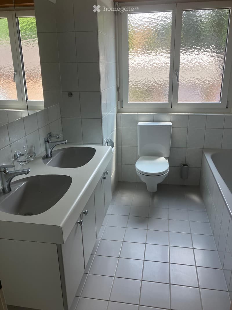 Modern bathroom with white tiles, a double sink, a toilet, and a bathtub. The room features large windows with frosted glass and a view of greenery outside.