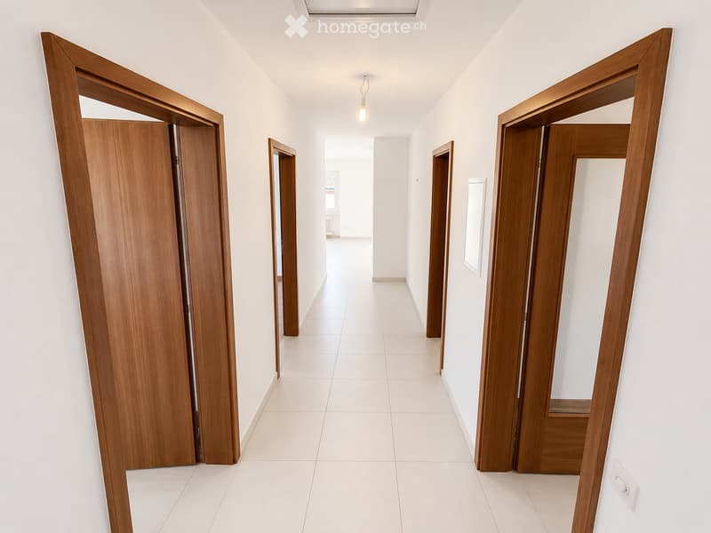 A clean, modern hallway with white tiled flooring, white walls, and several wooden doors. The hallway is well-lit with a single ceiling light fixture.