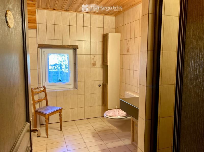 Bathroom with tiled walls and floor, a toilet, a small window, and a wooden chair with a blue seat.