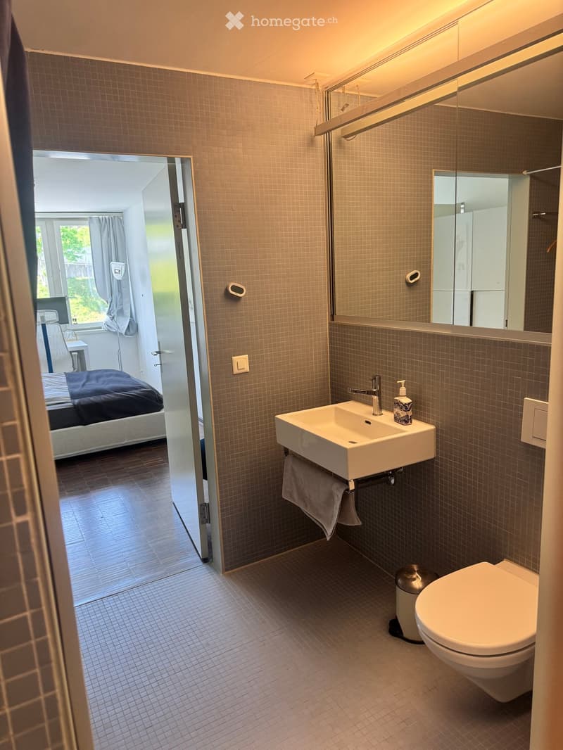 Modern bathroom with a white sink, toilet, large mirror, and towel. The adjacent bedroom is visible through an open door, featuring a bed with dark bedding and a window with curtains.