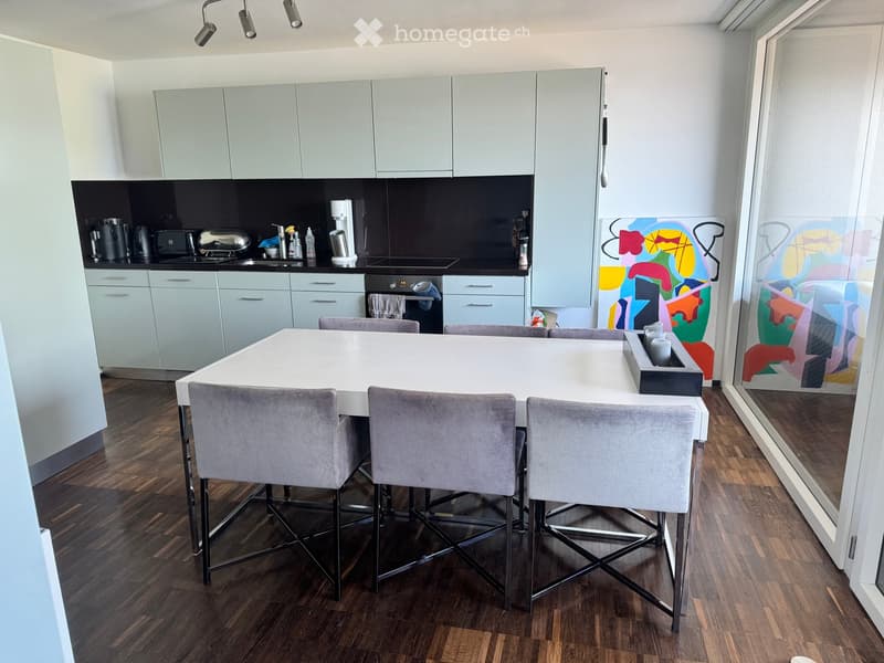 Modern kitchen with light grey cabinets, a white dining table with four grey chairs, and various appliances including a coffee maker, toaster, and oven. The floor is wooden, and there is a colorful abstract painting on the wall.