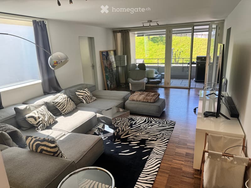 Modern living room with grey sectional sofa, zebra-patterned cushions, coffee table, ottoman, armchair, floor lamp, TV, and balcony access.
