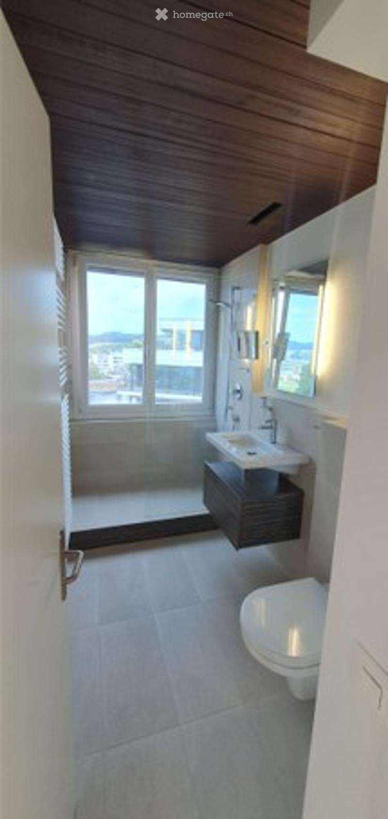 Modern bathroom with white tiles, wooden ceiling, large window, sink, toilet, and mirrored cabinet.