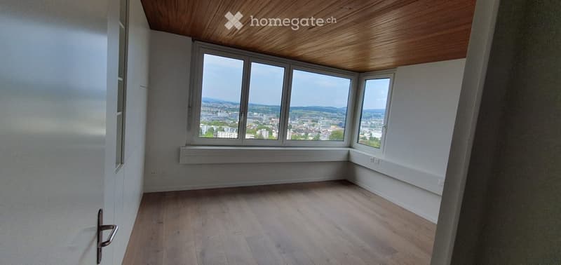 Empty room with wooden flooring, white walls, large windows offering a cityscape view, and a wooden slanted ceiling.