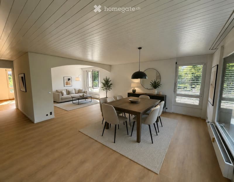 Open-concept living and dining area with light wood flooring, a beige sofa, coffee table, dining table with chairs, and modern decor including a large mirror and pendant light.