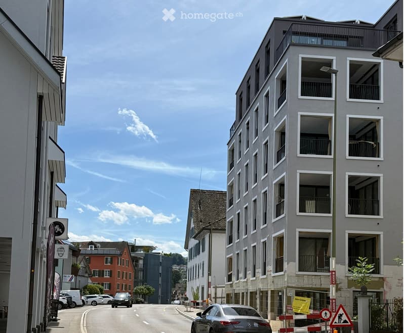 Modern multi-story apartment building with large windows and balconies, located on a quiet street with other buildings and parked cars visible.