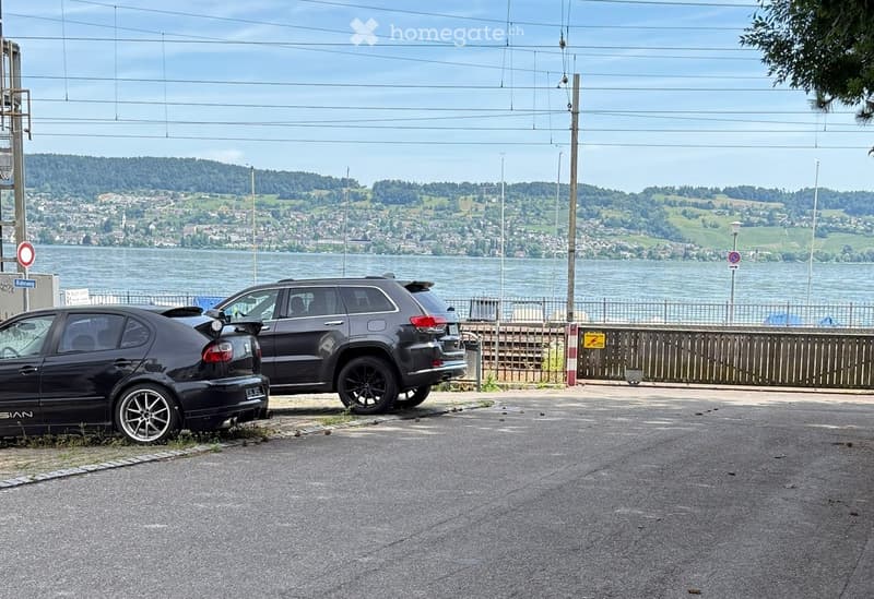 Two parked cars on a paved road near a waterfront with a scenic view of a town and hills across the water.