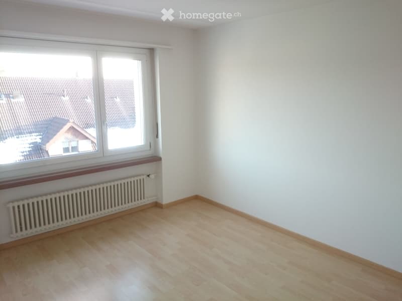Empty room with wooden flooring, white walls, large window with radiator below, and view of neighboring rooftops.