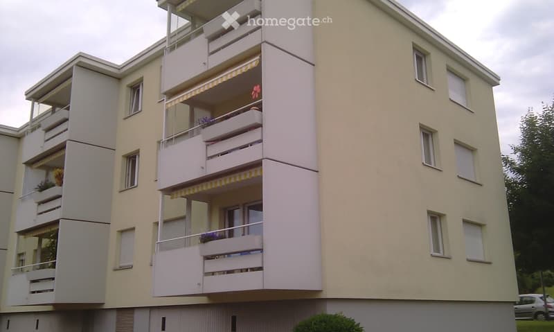 Multi-story residential building with multiple balconies, white railings, and flower boxes. Beige exterior walls and white-framed windows.