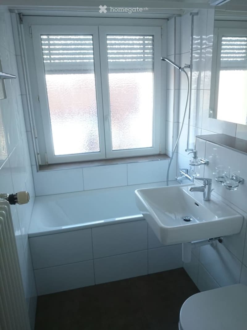 Modern bathroom with white tiles, a bathtub, a sink with a chrome faucet, a toilet, a radiator, and a large window with blinds.