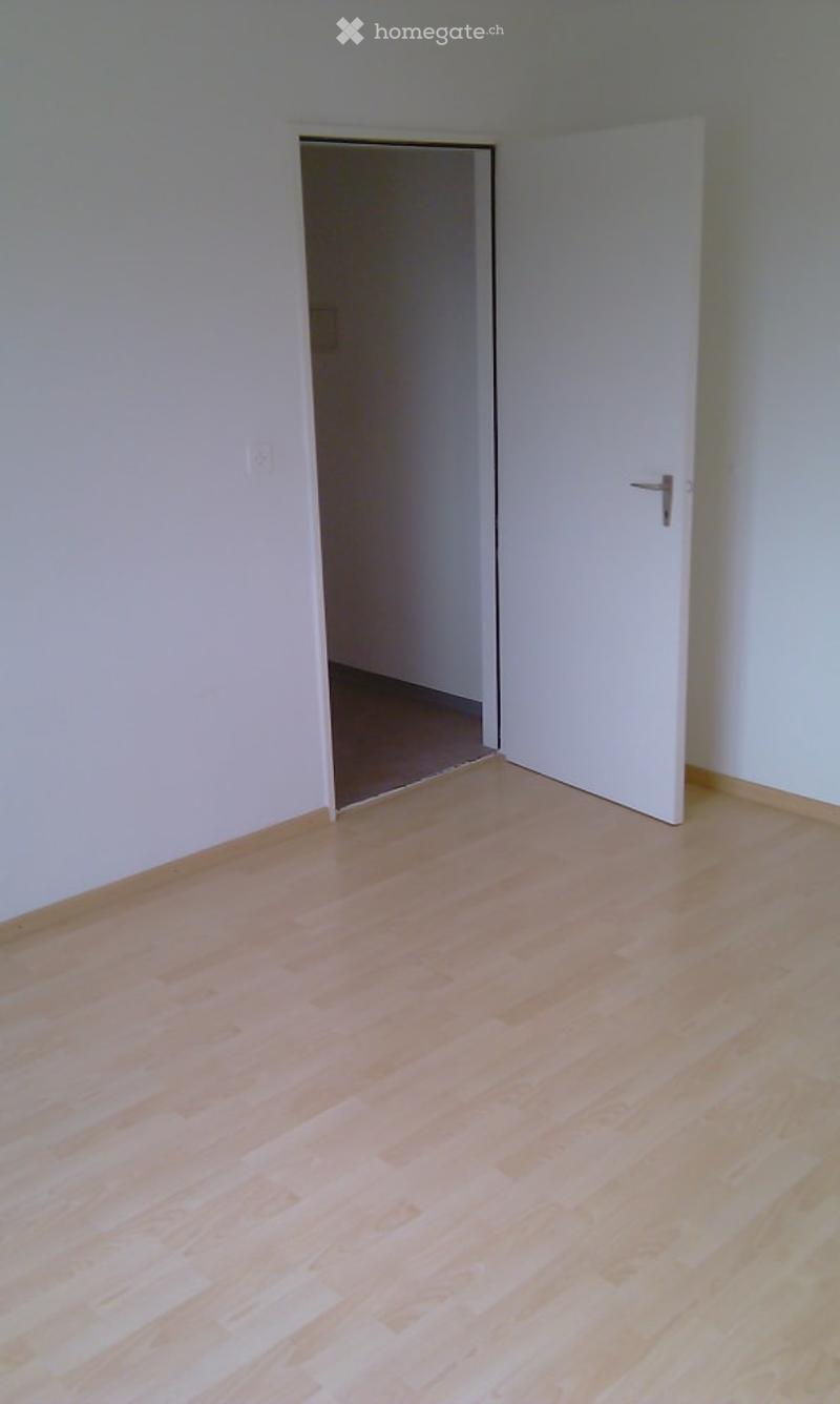 Empty room with light wooden flooring and white walls, featuring an open doorway leading to another room.