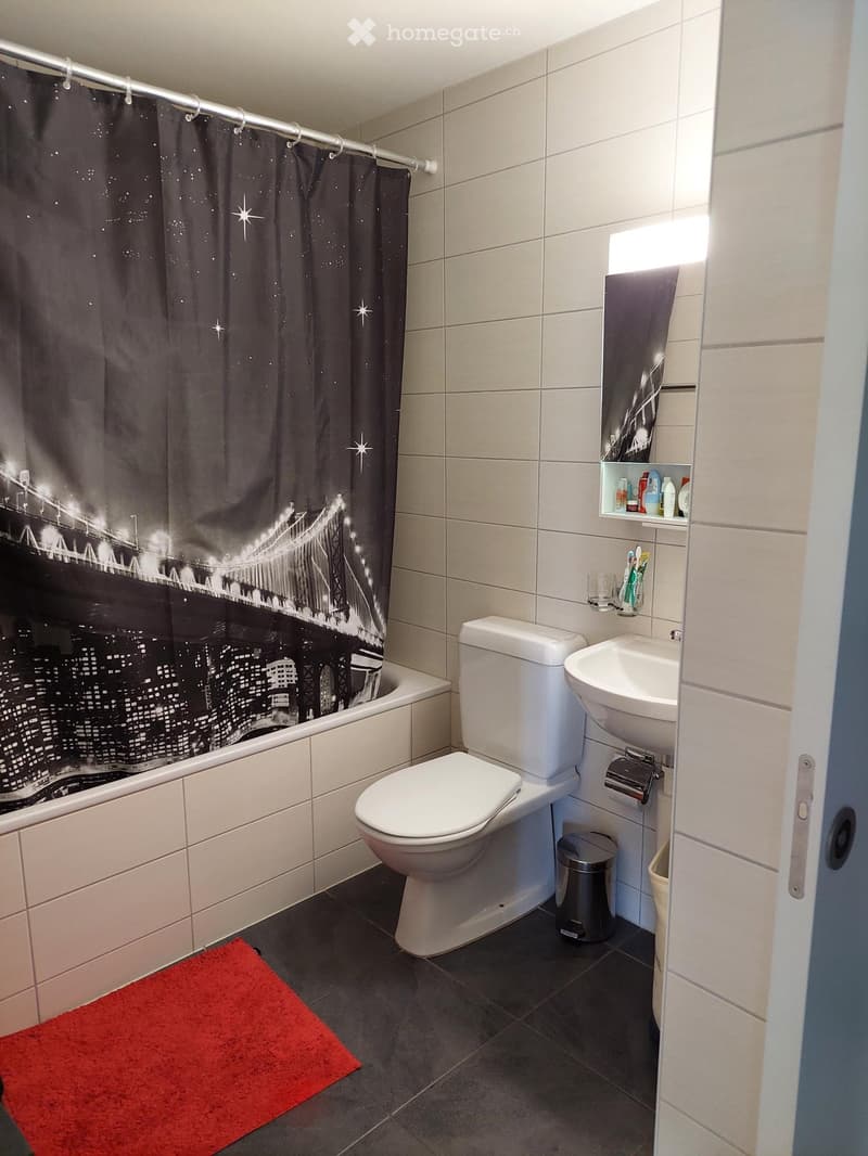 Modern bathroom with white tiles, a bathtub, toilet, sink, and a cityscape-themed shower curtain. Includes a small shelf with toiletries and a red bath mat.