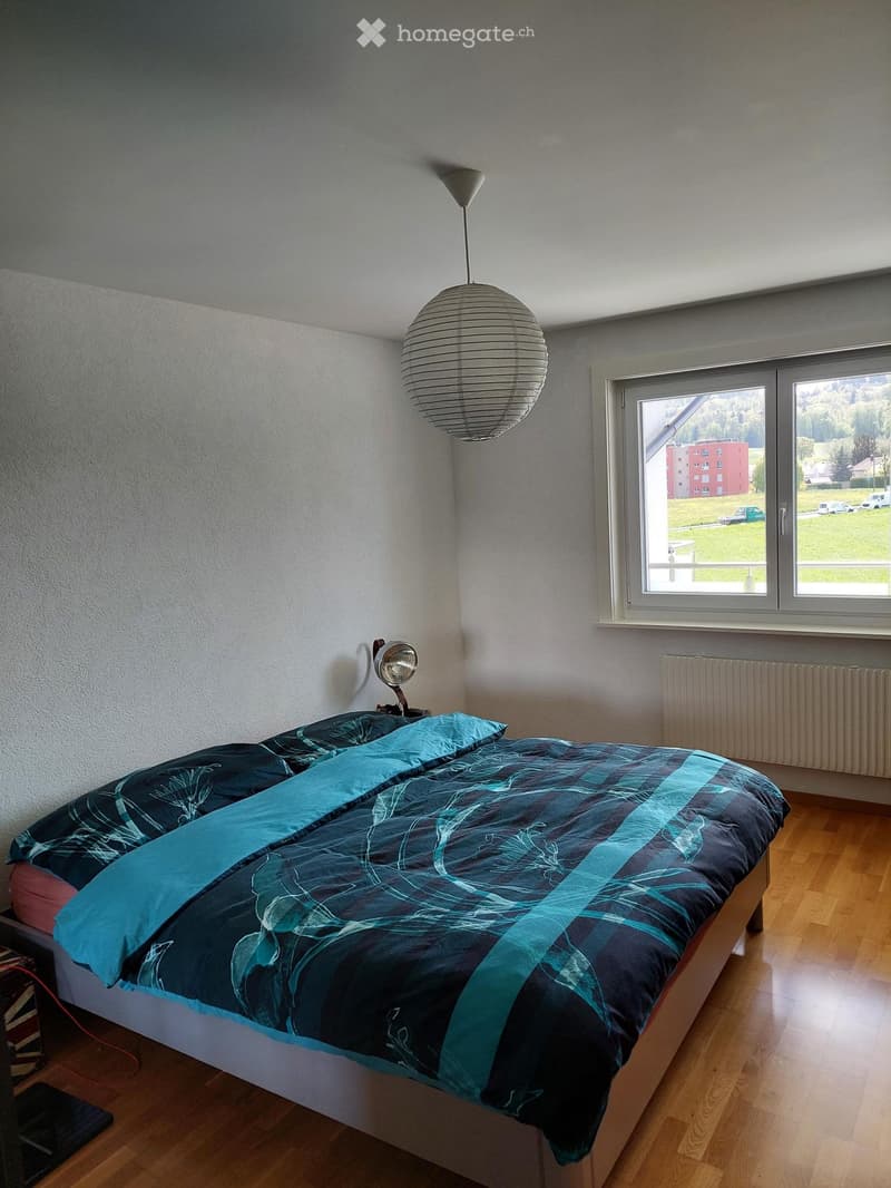 A modern bedroom with a wooden floor, a bed with a blue and black patterned bedspread, a wall-mounted lamp, and a large window offering a view of the landscape.