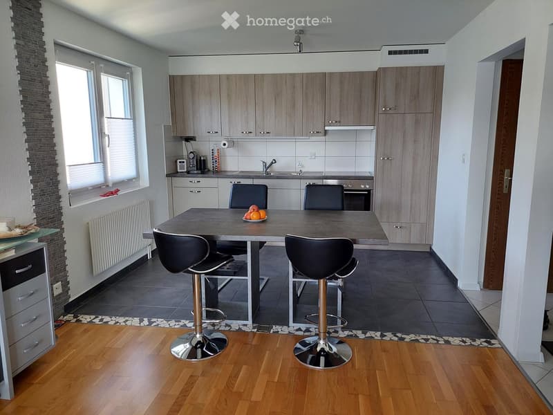 Modern kitchen with light wood cabinets, stainless steel appliances, a central island with bar stools, and a dining area.