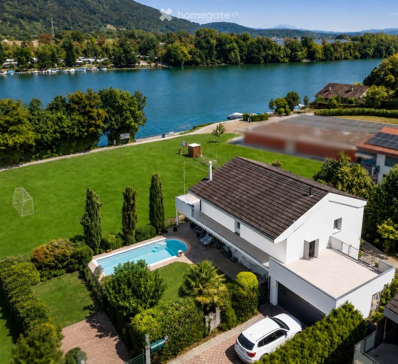 A modern two-story house with a dark roof and white walls, situated near a lake with a swimming pool, lawn, and surrounding greenery.