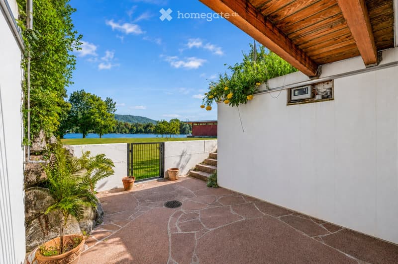 A serene outdoor patio with a stone-tiled floor, potted plants, a small staircase leading to a gated area, and a scenic view of a lake and distant hills.