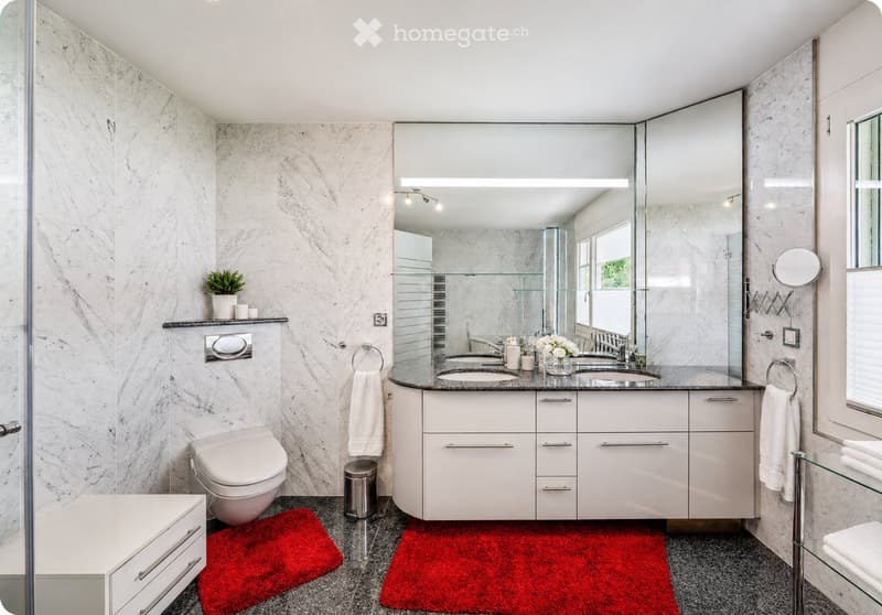 Modern bathroom with marble walls and floors, double sink vanity with white cabinets, large mirror, toilet, and decorative plant on shelf.