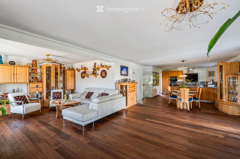 Spacious living and dining area with hardwood flooring, white sofa, coffee table, ottoman, dining table with chairs, kitchen area with cabinets and appliances, and decorative shelves.