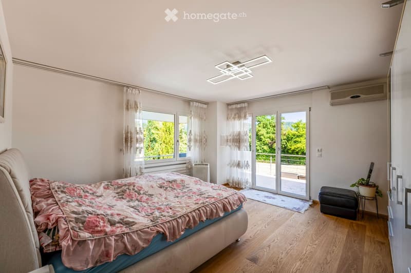 Bright bedroom with wooden flooring, large windows, and a sliding glass door leading to a balcony. Features a bed with floral bedding, a small black stool, and a potted plant near the door.
