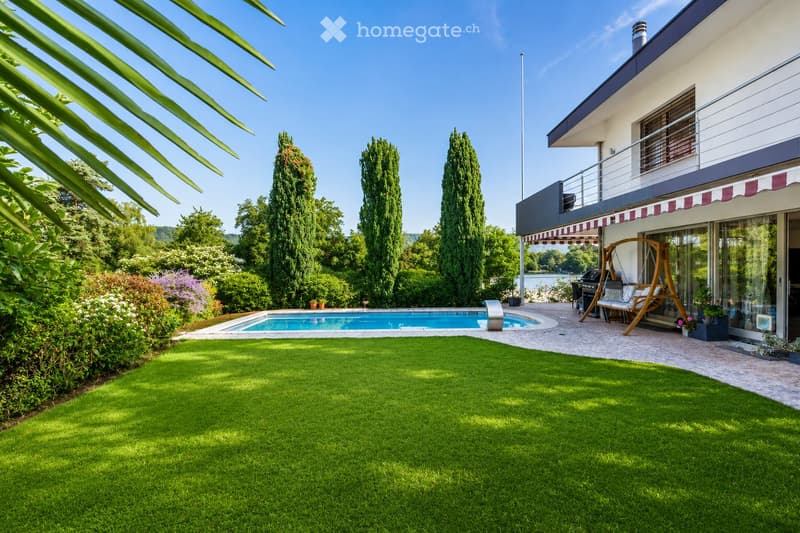 Well-maintained garden with a swimming pool, green lawn, tall trees, and a modern house with a balcony and outdoor seating area.