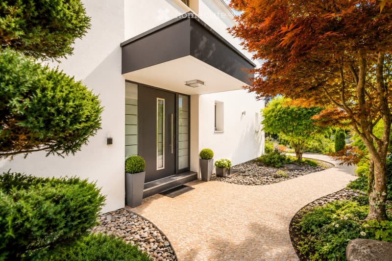 Modern two-story house with white exterior, black roof, and a small porch area. The entrance features a dark door with glass panels, flanked by small potted plants. The garden includes neatly trimmed bushes, a tree with red leaves, and a stone pathway lea