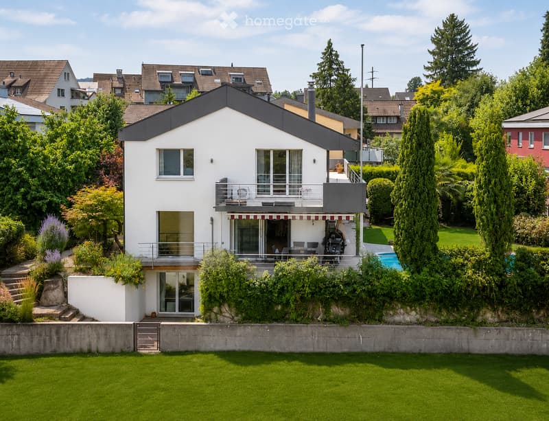 A modern two-story house with white exterior, large windows, and a balcony. The house features a well-maintained garden with green lawns, trees, and a swimming pool in the background.