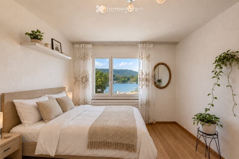Cozy bedroom with a large window overlooking a lake, featuring a wooden bed, nightstand, mirror, and potted plants.
